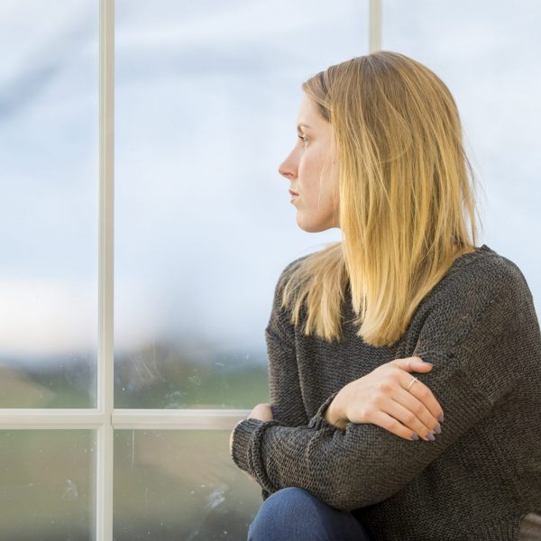 A caucasian woman in her 20s sits at the window and gazes outside. She has her arms crossed and is alone.